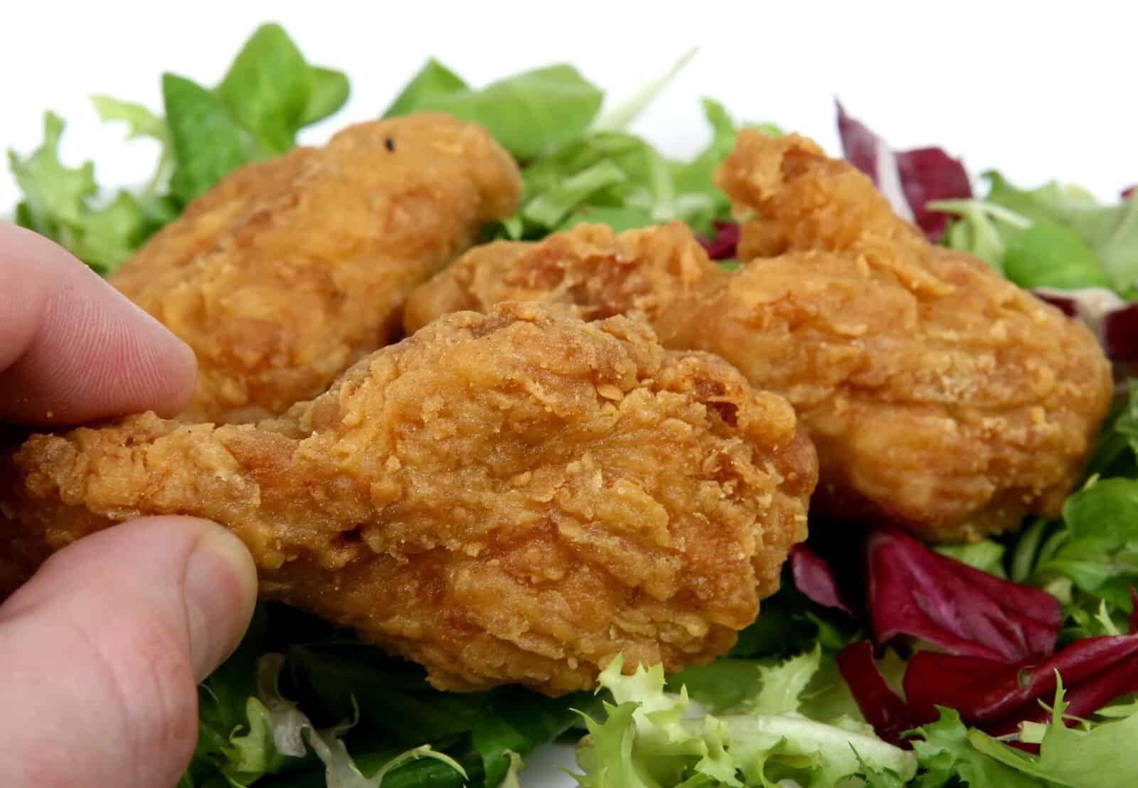 fried chicken leg held by fingers of a hand on a bed of green lettuce
